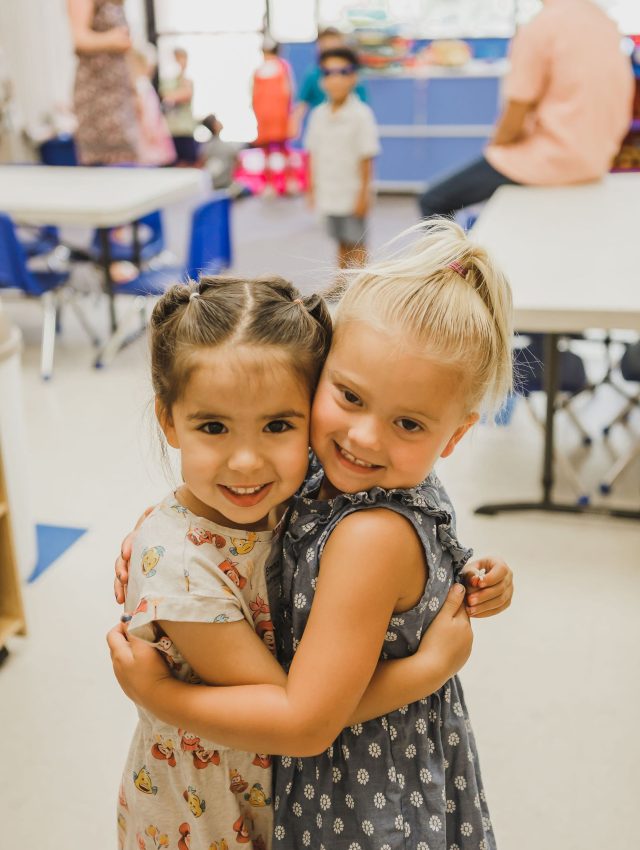 Two young girls hugging and smiling at the camera in a Faith Community Church classroom